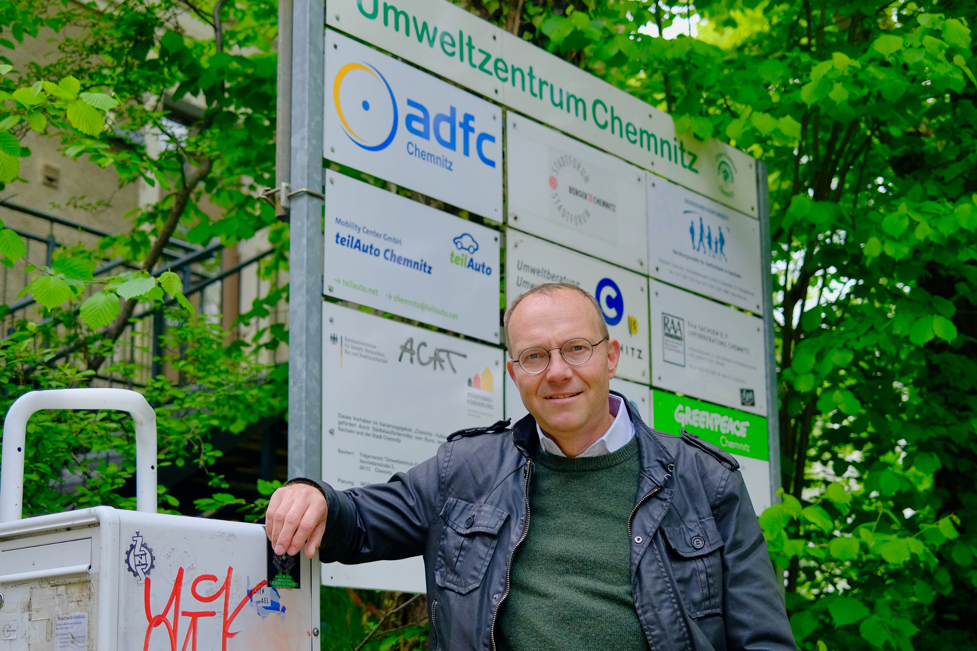 Wolfram Günther vor dem Umweltzentrum Chemnitz, zu sehen ist eine Tafel mit den hier ansässigen Vereinen, Initiativen und Unternehmen.